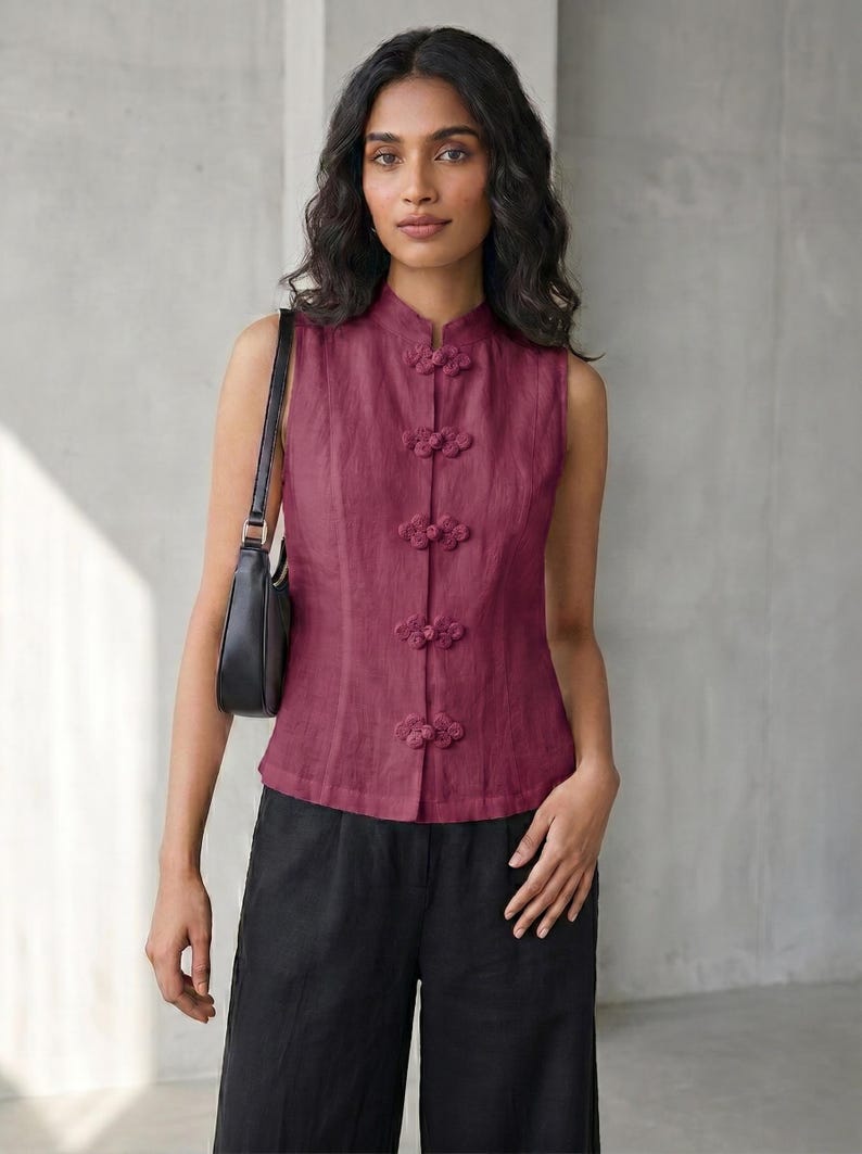 May include: A sleeveless, burgundy top with a mandarin collar and decorative frog closures. The top is paired with black pants and a black shoulder bag. The outfit is simple and elegant.
