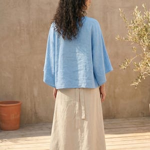 May include: A light blue linen top with wide sleeves and a beige linen skirt. The skirt has a wrap-around design with a matching belt. The outfit is worn outdoors on a wooden deck.