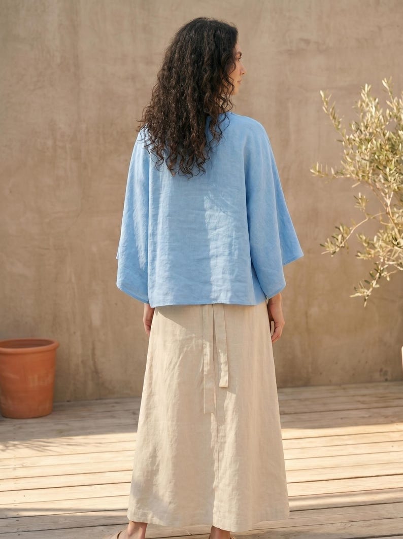 May include: A light blue linen top with wide sleeves and a beige linen skirt. The skirt has a wrap-around design with a matching belt. The outfit is worn outdoors on a wooden deck.