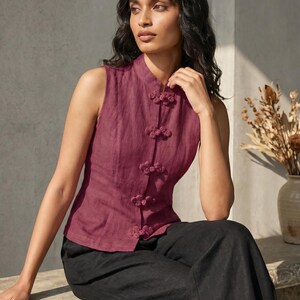 May include: A sleeveless, burgundy top with a mandarin collar and decorative frog closures. The top is paired with black pants. A vase with dried flowers is in the background.