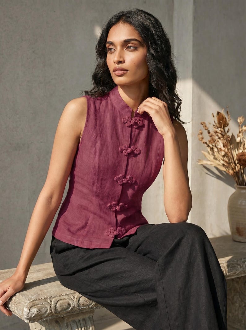 May include: A sleeveless, burgundy top with a mandarin collar and decorative frog closures. The top is paired with black pants. A vase with dried flowers is in the background.