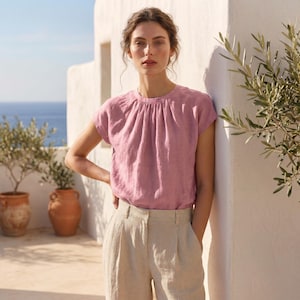 Peut inclure: Un haut rose poudr&eacute; &agrave; manches courtes avec un col fronc&eacute;. Le haut est associ&eacute; &agrave; un pantalon en lin beige clair. L'arri&egrave;re-plan pr&eacute;sente un mur blanc, des pots en terre cuite et une vue sur la mer.