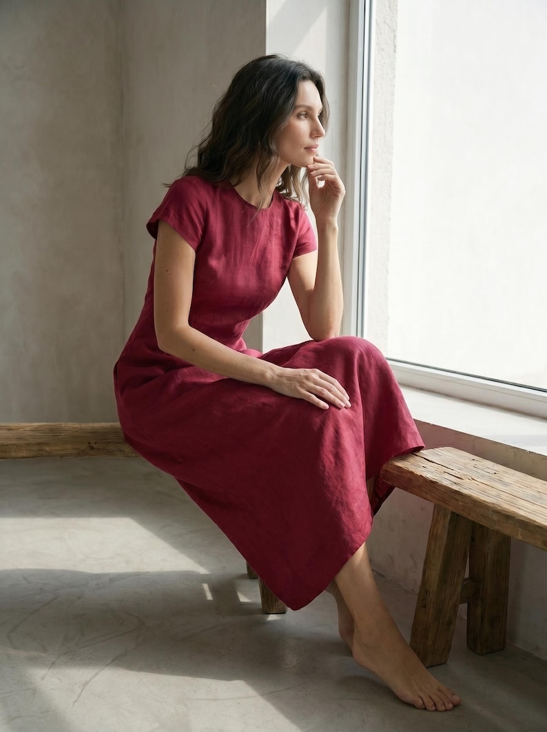 May include: A burgundy-colored linen dress with short sleeves and a round neckline. The dress is knee-length and worn by a person sitting near a window. The person is sitting on a wooden bench.