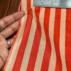 Op de afbeelding: Close-up van een stof met verticale strepen in afwisselende tinten rood en beige. De stof wordt vastgehouden en een metalen liniaal is zichtbaar, wat de breedte van het materiaal aangeeft. De stof lijkt lichtgewicht en mogelijk van linnen of katoen.