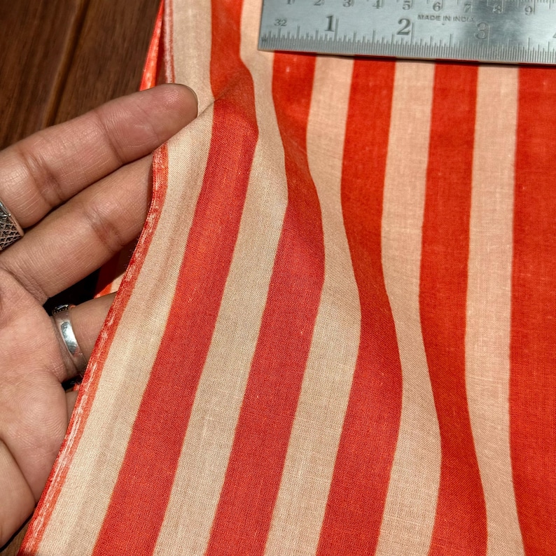 Op de afbeelding: Close-up van een stof met verticale strepen in afwisselende tinten rood en beige. De stof wordt vastgehouden en een metalen liniaal is zichtbaar, wat de breedte van het materiaal aangeeft. De stof lijkt lichtgewicht en mogelijk van linnen of katoen.