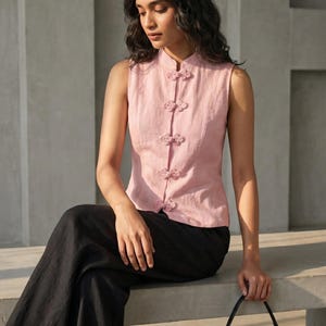 May include: A sleeveless, light pink top with a mandarin collar and decorative floral frog closures. The top is paired with black pants. A black handbag is held in the hand.
