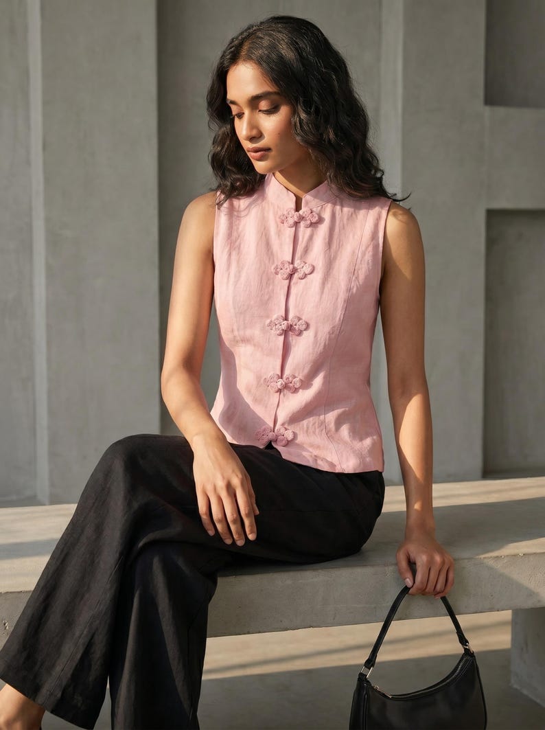 May include: A sleeveless, light pink top with a mandarin collar and decorative floral frog closures. The top is paired with black pants. A black handbag is held in the hand.
