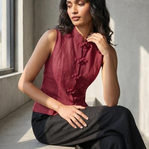 May include: A sleeveless burgundy top with a mandarin collar and decorative knot closures. The top is paired with black pants. The outfit is made of a lightweight fabric, suitable for warm weather. The style is reminiscent of traditional Asian designs.