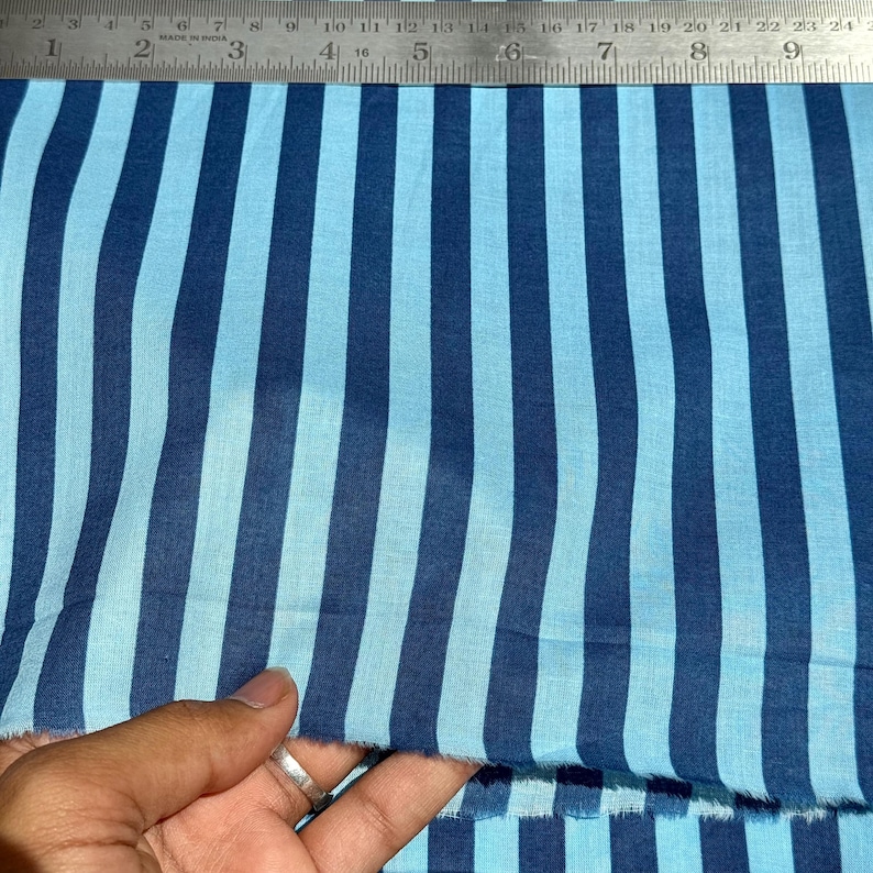 Op de afbeelding: Blauw gestreepte stof met afwisselend licht- en donkerblauwe verticale strepen. Een metalen liniaal met centimeter maten ligt bovenop. De stof wordt door een hand vastgehouden. Het patroon van de stof doet denken aan een nautisch thema.