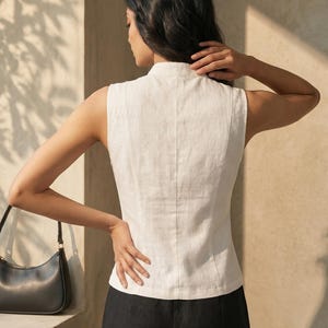 May include: Sleeveless white linen top with a high collar and vertical seams down the back. The top is paired with black pants. A black purse hangs on the left side. The outfit is simple and elegant.