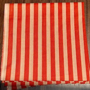 Op de afbeelding: Een gevouwen textiel met een opvallend streepjespatroon. Het ontwerp heeft verticale strepen in afwisselende banden van felrood en cr&egrave;me. De stof lijkt licht van gewicht, mogelijk linnen of katoen, en is gevouwen.