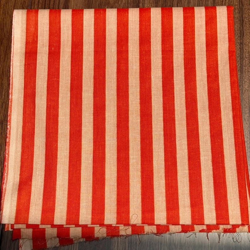 Op de afbeelding: Een gevouwen textiel met een opvallend streepjespatroon. Het ontwerp heeft verticale strepen in afwisselende banden van felrood en cr&egrave;me. De stof lijkt licht van gewicht, mogelijk linnen of katoen, en is gevouwen.