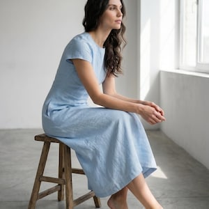 May include: A light blue linen dress with short sleeves. The dress is worn by a person sitting on a wooden stool. The person has long dark hair and is looking out a window. The dress has a simple, elegant design.