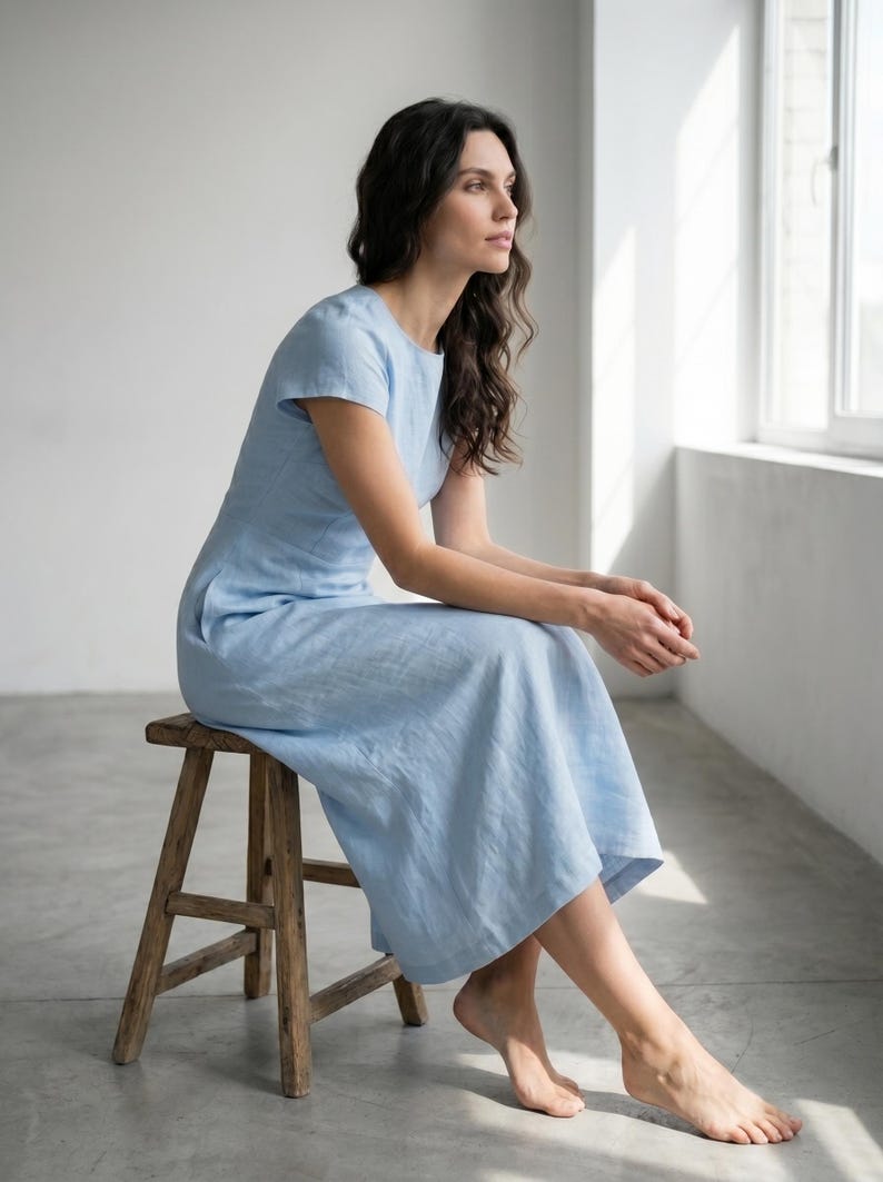 May include: A light blue linen dress with short sleeves. The dress is worn by a person sitting on a wooden stool. The person has long dark hair and is looking out a window. The dress has a simple, elegant design.