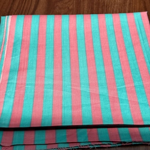 Op de afbeelding: Een gevouwen textiel met een streepjespatroon. De verticale strepen wisselen af in turquoise en koraalroze. De stof lijkt licht, mogelijk katoen of linnen. De kleuren zijn helder en het patroon is regelmatig.
