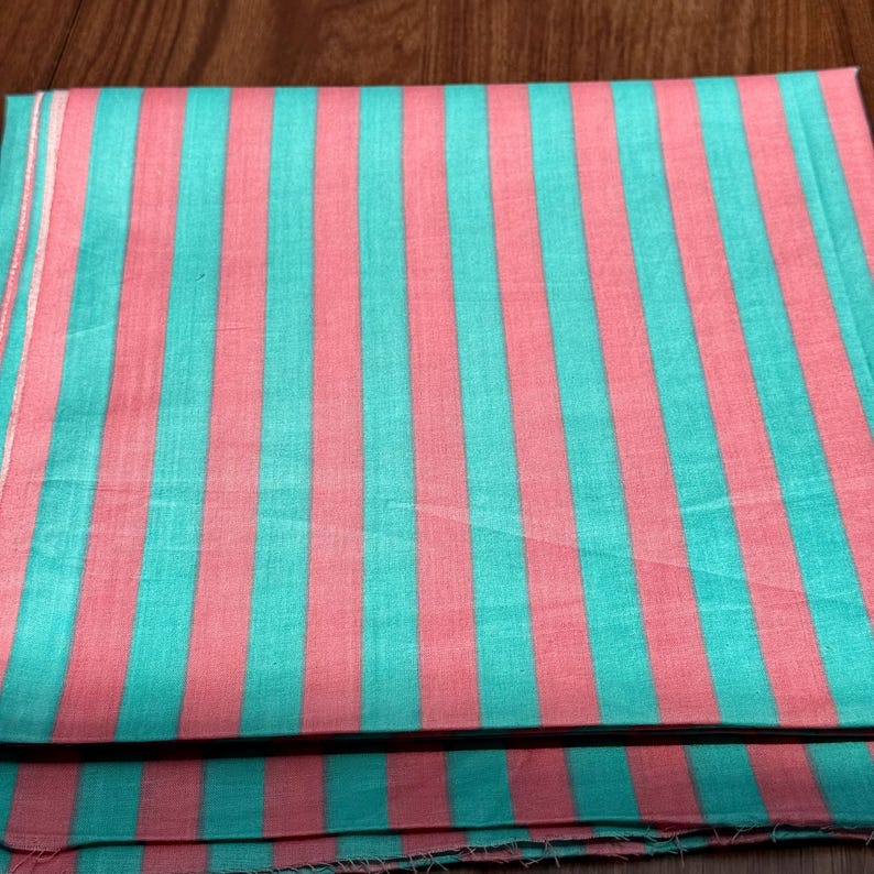 Op de afbeelding: Een gevouwen textiel met een streepjespatroon. De verticale strepen wisselen af in turquoise en koraalroze. De stof lijkt licht, mogelijk katoen of linnen. De kleuren zijn helder en het patroon is regelmatig.
