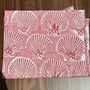 Pode incluir: Tecido dobrado com um padrão repetido de conchas brancas sobre um fundo rosa coral. As conchas são detalhadas com linhas verticais, criando uma aparência texturizada. O tecido está cuidadosamente dobrado, mostrando o design.