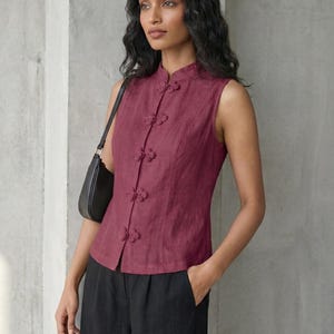 May include: A sleeveless burgundy top with a mandarin collar and decorative frog closures. The top is paired with black pants and a black shoulder bag. The style is reminiscent of traditional Asian designs.