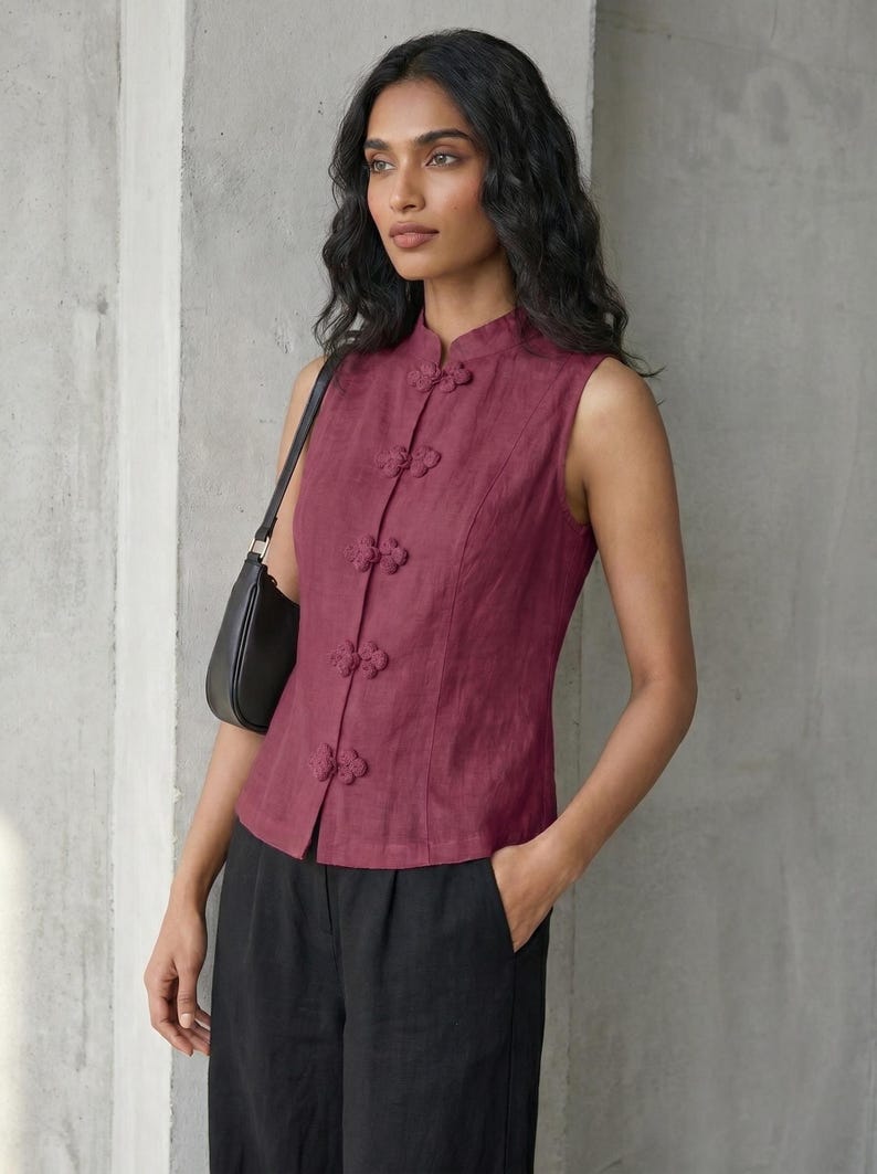 May include: A sleeveless burgundy top with a mandarin collar and decorative frog closures. The top is paired with black pants and a black shoulder bag. The style is reminiscent of traditional Asian designs.