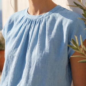 Peut inclure: Un haut en lin bleu clair avec un col rond et des manches courtes. Le haut pr&eacute;sente des d&eacute;tails fronc&eacute;s sur la poitrine. Le tissu a un aspect l&eacute;g&egrave;rement textur&eacute;. Le v&ecirc;tement est de couleur unie.
