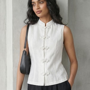 May include: White sleeveless top with a mandarin collar and floral frog closures. The top is made of a textured fabric and is paired with black pants. A black shoulder bag is also visible.
