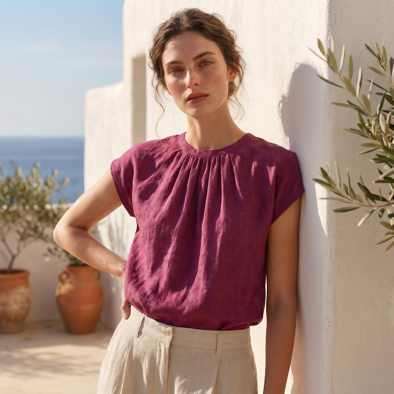 Peut inclure: Un haut en lin magenta avec un col fronc&eacute; et des manches courtes. Le haut est associ&eacute; &agrave; un pantalon en lin beige. Le mod&egrave;le est appuy&eacute; contre un mur blanc, avec la mer et des oliviers en arri&egrave;re-plan.