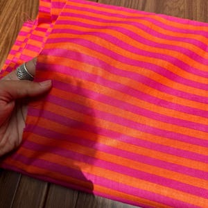 Op de afbeelding: Een levendige stof met afwisselende horizontale strepen in felroze en oranje. De stof is gevouwen en toont de textuur en glans van het materiaal. Een hand met een zilveren ring houdt de stof vast.