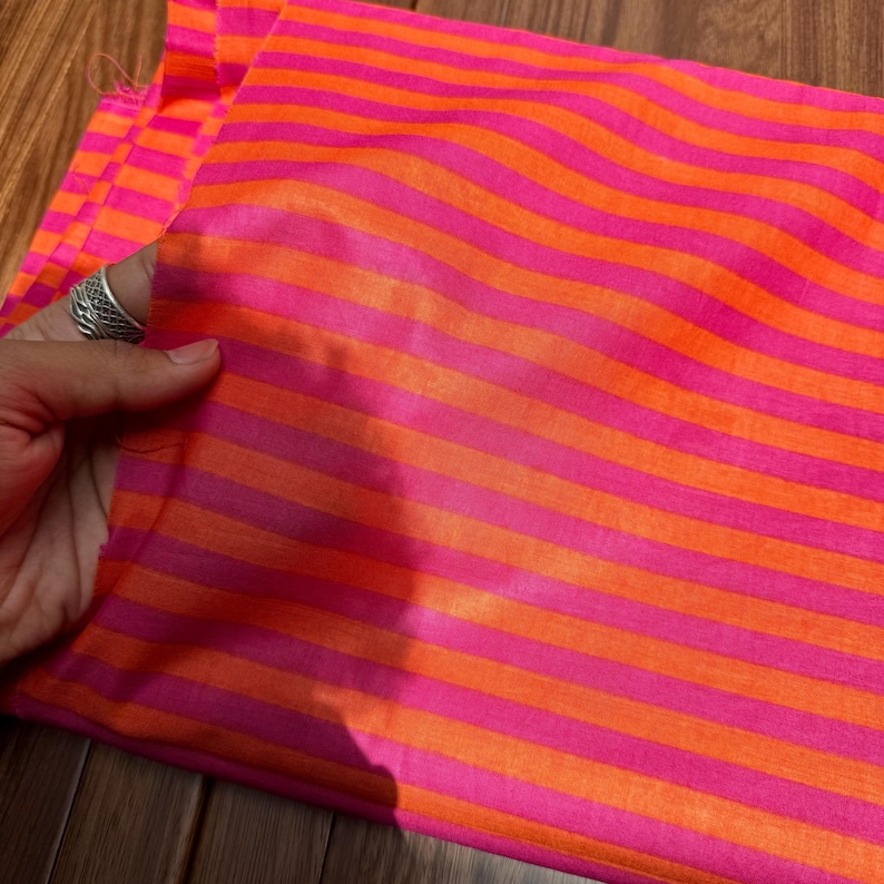 Op de afbeelding: Een levendige stof met afwisselende horizontale strepen in felroze en oranje. De stof is gevouwen en toont de textuur en glans van het materiaal. Een hand met een zilveren ring houdt de stof vast.