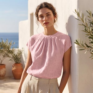May include: A light pink linen top with short sleeves and a gathered neckline. The top is paired with beige linen pants. The model is leaning against a white wall with olive trees in the background, creating a soft, natural aesthetic.