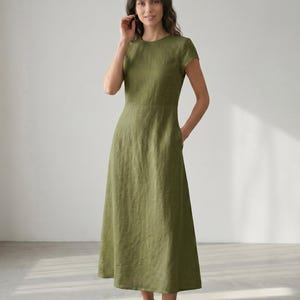 May include: A long olive green dress with short sleeves and a round neckline. The dress has a fitted bodice and a flared skirt, with pockets on each side. The model is standing in a well-lit room.