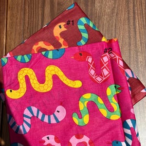 Op de afbeelding: Twee kleurrijke stoffen met slangenpatronen. Eén is dieprood met oranje, blauwe en groene slangen, gemarkeerd met "#1". De andere is felroze met gele, groene en roze slangen, gemarkeerd met "#2".