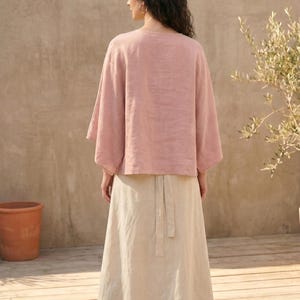 May include: A woman wearing a light pink linen top with wide sleeves and a long, beige skirt. The outfit is simple and elegant, suitable for warm weather. The background includes a wooden deck, a terracotta pot, and a plant.