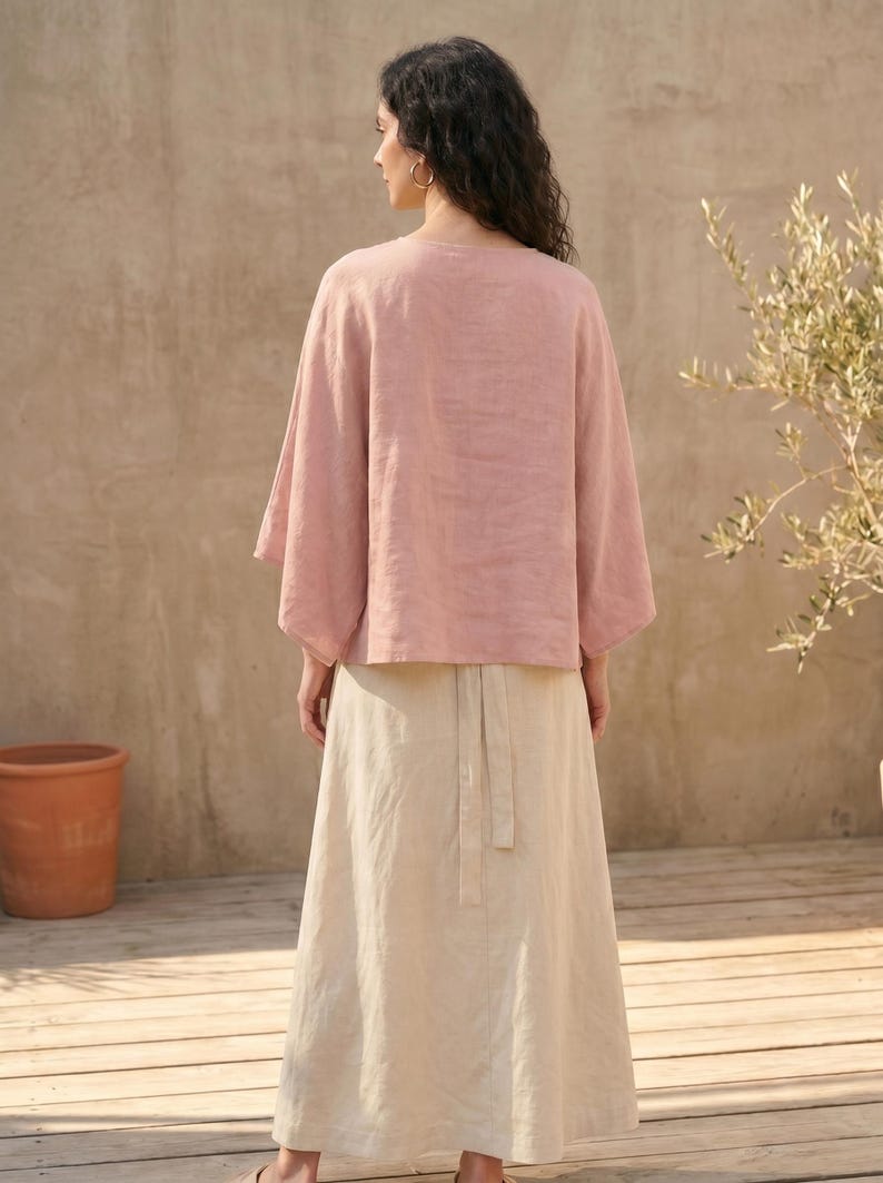 May include: A woman wearing a light pink linen top with wide sleeves and a long, beige skirt. The outfit is simple and elegant, suitable for warm weather. The background includes a wooden deck, a terracotta pot, and a plant.