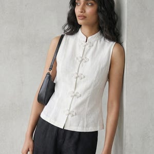 May include: A sleeveless white top with a mandarin collar and decorative floral-shaped buttons. The top is paired with black pants and a black shoulder bag. The outfit is simple and elegant.
