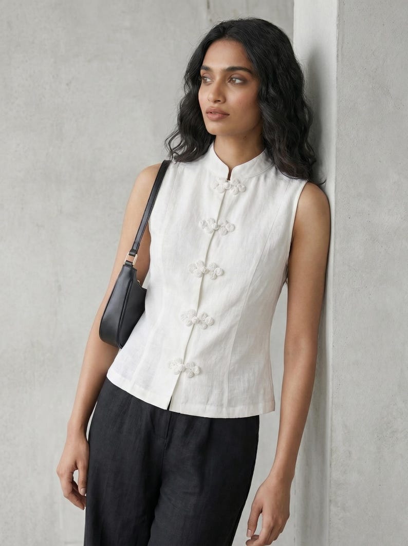 May include: A sleeveless white top with a mandarin collar and decorative floral-shaped buttons. The top is paired with black pants and a black shoulder bag. The outfit is simple and elegant.