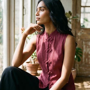 May include: A sleeveless, burgundy top with a mandarin collar and decorative knot closures. The top is paired with black pants. The woman is sitting near a window, with plants in the background.