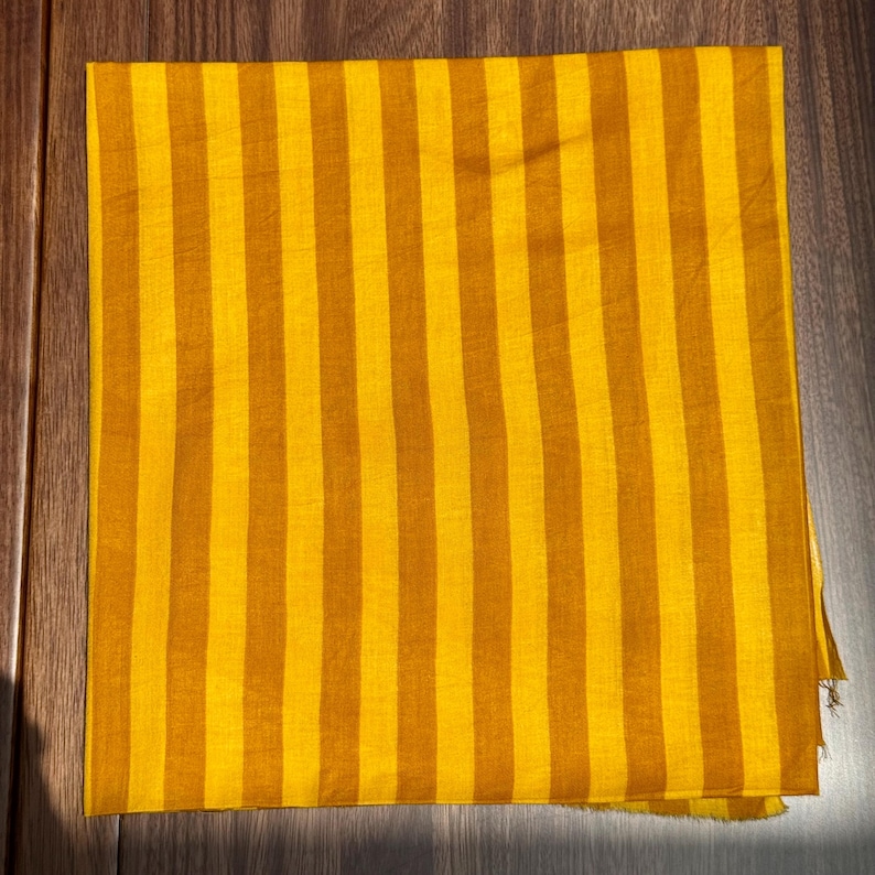 Op de afbeelding: Een gevouwen textiel met verticale strepen in tinten geel en bruin. De stof is rechthoekig en lijkt gemaakt van een lichtgewicht materiaal. De strepen zijn gelijkmatig verdeeld en lopen over de lengte van de stof. Het textiel is waarschijnlijk een sjaal of een vergelijkbaar accessoire.