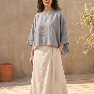May include: A woman wearing a gray linen top with wide sleeves and a long, cream-colored linen skirt. The outfit is simple and elegant, suitable for warm weather. The background features a wooden deck and a neutral-toned wall.