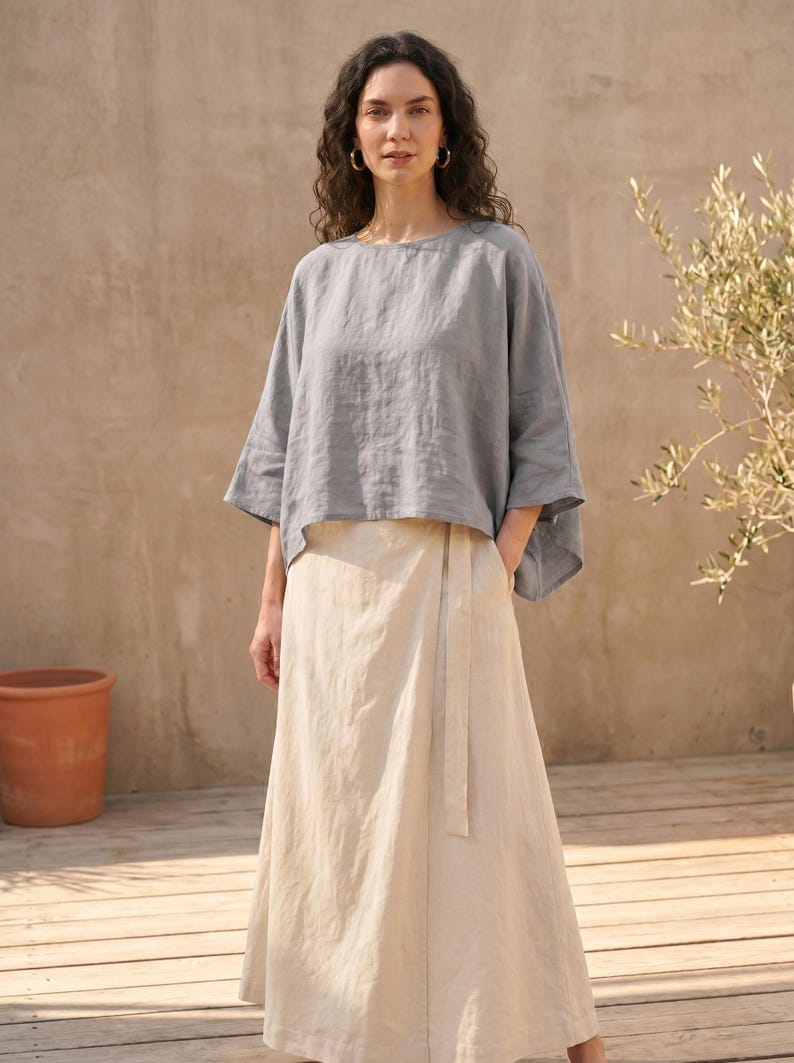 May include: A woman wearing a gray linen top with wide sleeves and a long, cream-colored linen skirt. The outfit is simple and elegant, suitable for warm weather. The background features a wooden deck and a neutral-toned wall.