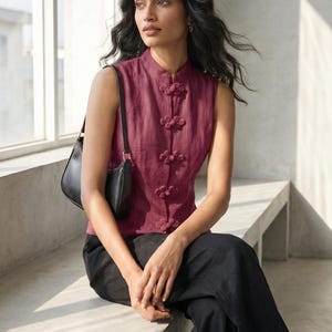 May include: A sleeveless burgundy top with a mandarin collar and decorative frog closures. The top is paired with black pants and a black shoulder bag. The woman is sitting on a concrete bench near a window.