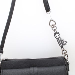May include: Black crossbody bag with a textured finish, featuring a decorative chain with heart-shaped links and a charm of a skeleton cat. The bag has a long, adjustable strap and silver-tone hardware.