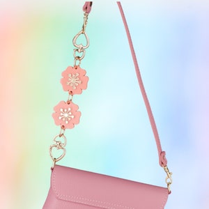 May include: A pink leather crossbody bag with a decorative chain strap. The chain features gold-colored heart-shaped links and pink flower charms. The bag has a matching pink strap and gold-colored hardware.