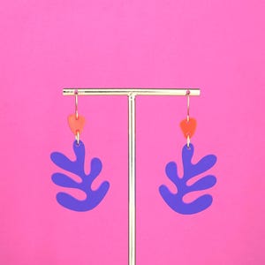 May include: A pair of earrings with a gold-coloured hook, an orange heart-shaped accent, and a large, leaf-shaped, purple pendant. The earrings are displayed against a bright pink background.