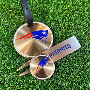 Könnte beinhalten: Ein goldfarbener Golfball-Marker mit einem rot-weiß-blauen Patriots-Logo. Ein passender goldfarbener Golfball-Retriever mit einem Patriots-Logo und einem schwarzen Lederband mit einer goldenen Schnalle. Die Worte "PATRIOTS" sind auf den Retriever gedruckt.