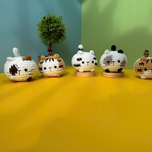 K&ouml;nnte beinhalten: F&uuml;nf geh&auml;kelte Katzenfiguren in verschiedenen Farben und Mustern. Jede Katze ist rund mit kleinen Ohren und einem Schwanz. Die Katzen sind wei&szlig;, braun und grau, mit schwarzen Gesichtsdetails. Sie sitzen auf kleinen Holzsockeln, die auf einer gelben Oberfl&auml;che ausgestellt sind.