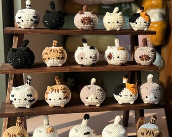 May include: A wooden shelf displays several handmade crochet cat figurines. The cats are round with small tails and various colour patterns, including black, white, brown, and calico. The shelf has three tiers and is made of dark wood.