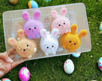 May include: A clear plastic container holds five crocheted bunny figures in various pastel colors: lavender, pink, white, yellow, and tan. The bunnies have black eyes and pink accents. Decorative Easter eggs in various colors are scattered around the container on a bed of green grass.