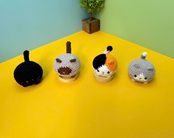 May include: Four handmade crochet cat-shaped bottle stoppers. The cats are black, grey, calico, and grey. Each cat has a tail and is sitting on a wooden base. The background is yellow, teal, and green.