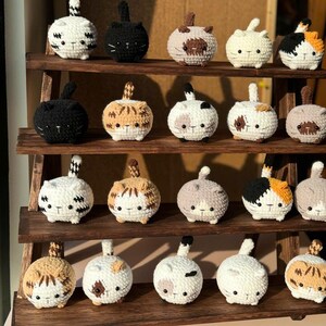 K&ouml;nnte beinhalten: Ein Holzregal zeigt eine Sammlung geh&auml;kelter Katzenfiguren. Die Katzen sind rund mit kleinen Schw&auml;nzen und verschiedenen Farbkombinationen, darunter Wei&szlig;, Schwarz, Braun und Schildpattmuster. Jede Katze hat ein gesticktes Gesicht mit kleinen Augen und einer Nase.