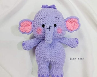 May include: A lilac crochet elephant toy with pink inner ears and rosy cheeks. The handmade elephant has a small trunk and a cute expression. The toy is soft and plush, with a bow on its head. The text "@Lan Tran" is visible.